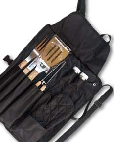 Homem sorrindo, em pé, segurando um kit de churrasco completo com avental preto e utensílios de metal, como faca, garfo e espátula. Ele veste uma jaqueta de couro e possui uma mochila nas costas.