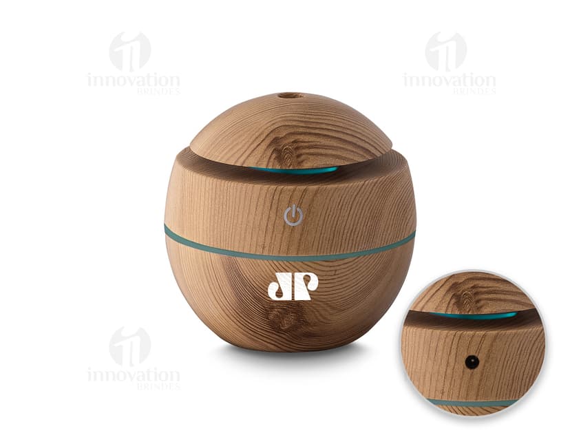Umidificador de ar USB em ABS reciclável, que aparece em uma cesta de vime marrom ao lado de um banco, comida, dinheiro e outros objetos. A imagem é de vista próxima, com foco no produto e nos detalhes dos materiais utilizados.