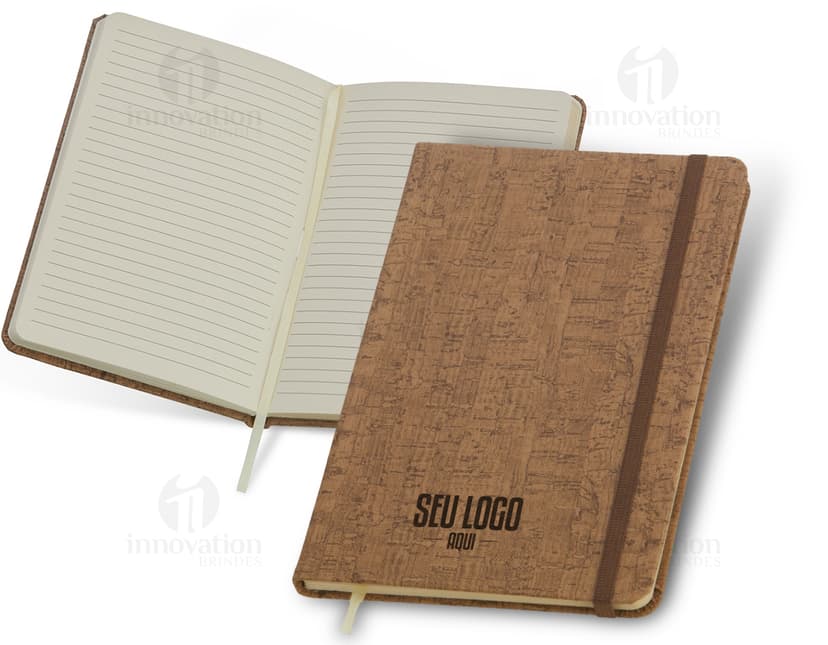 Caderneta em cortiça com marca página, em uma mesa de escritório vazia, ao lado de um notebook, um disco de vídeo, papel em branco e um livro aberto.
