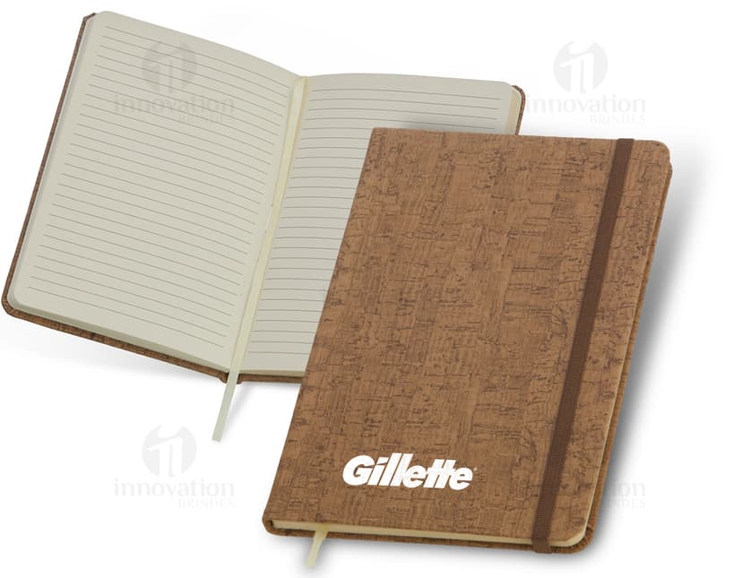 Caderneta em cortiça com marca página, em uma mesa de escritório vazia, ao lado de um notebook, um disco de vídeo, papel em branco e um livro aberto.