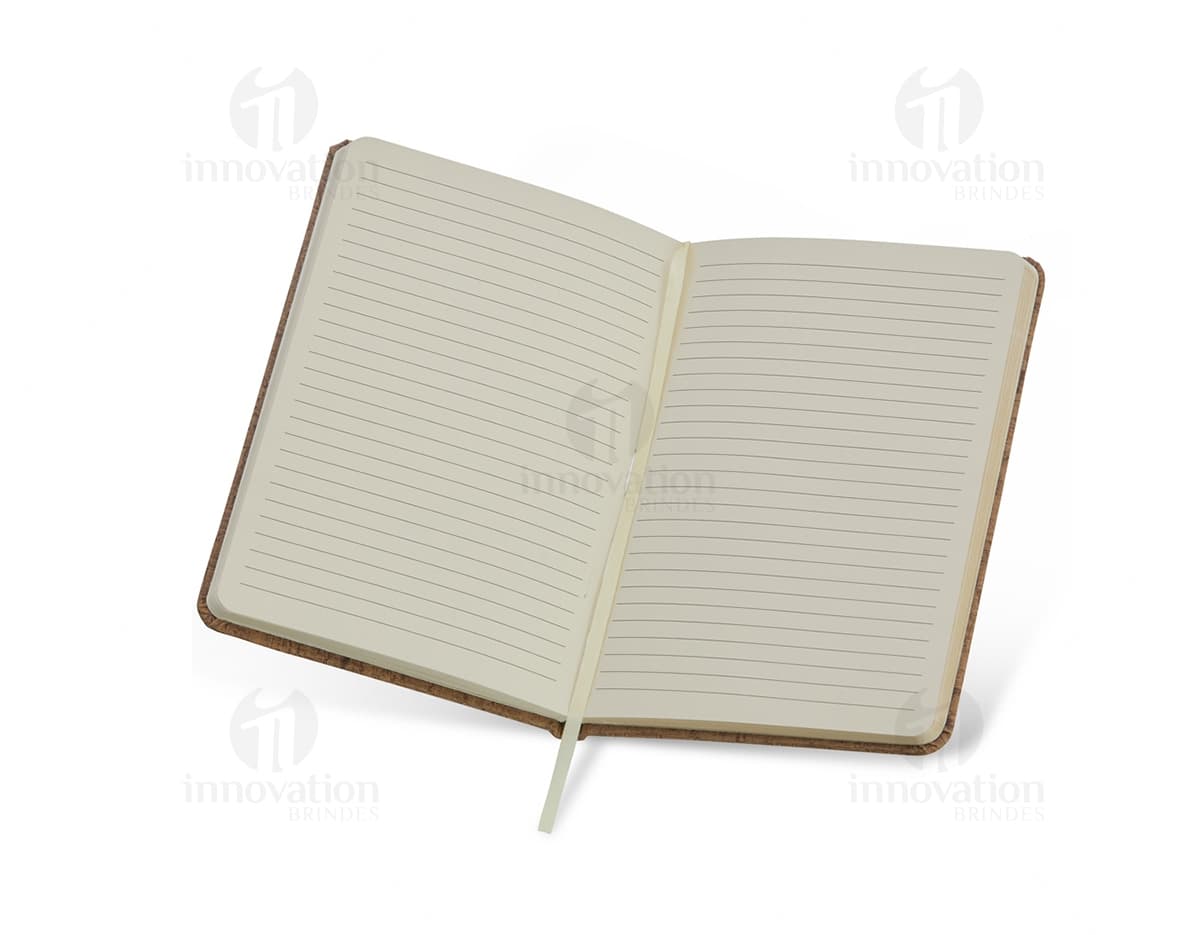 Caderneta em cortiça com marca página, em uma mesa de escritório vazia, ao lado de um notebook, um disco de vídeo, papel em branco e um livro aberto.