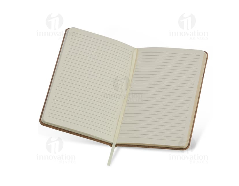Caderneta em cortiça com marca página, em uma mesa de escritório vazia, ao lado de um notebook, um disco de vídeo, papel em branco e um livro aberto.