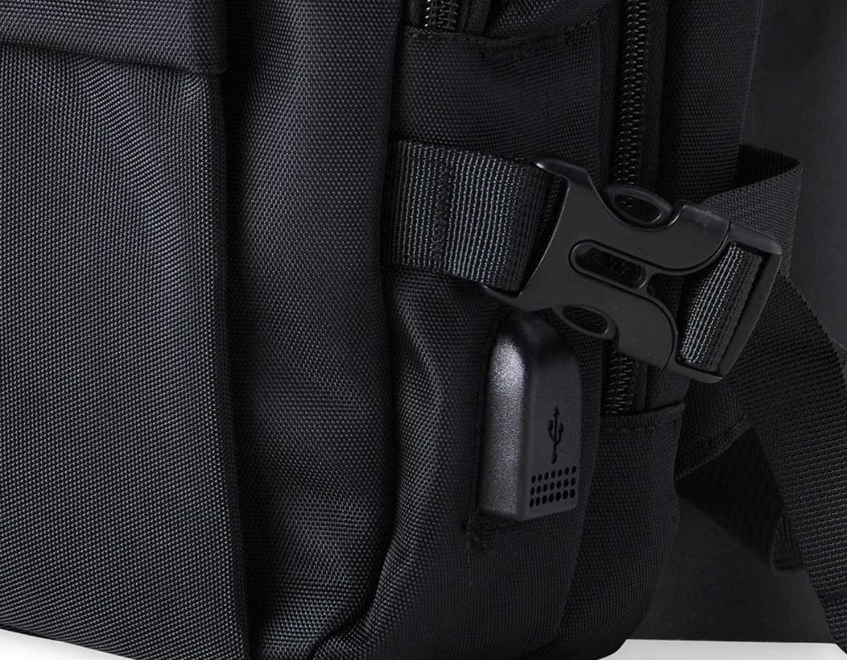 Homem de negócios usando um elegante terno preto, segurando uma mochila de nylon preta de 20 litros com USB. O acessório tem apoio nas costas, alça, e suporte para objetos.