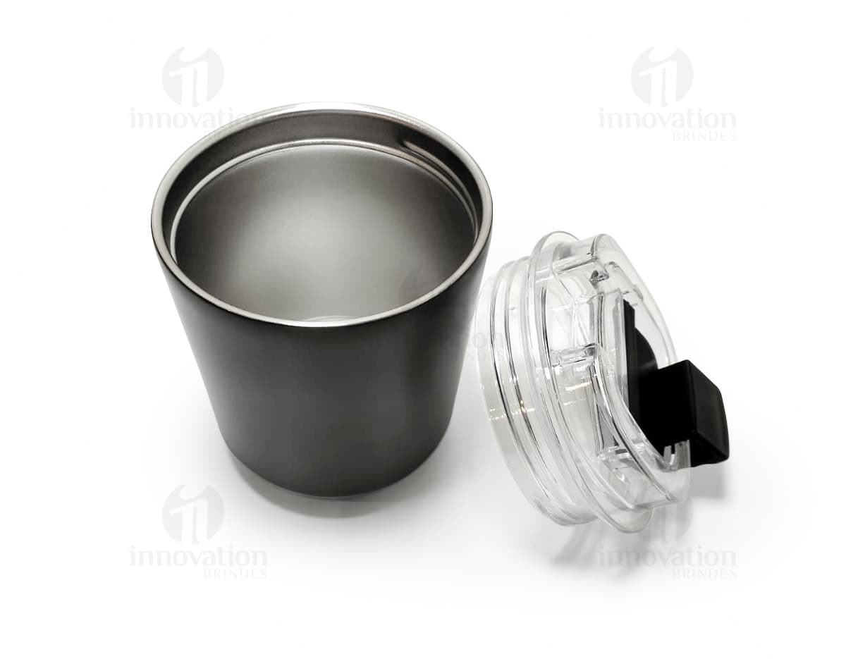 Copo térmico de 280ml em cerâmica preta e metal de cor prateada, com alça e tampa. O copo está vazio e posicionado sobre uma mesa de cozinha, ao lado de uma colher, uma jarra de café e alguns utensílios.