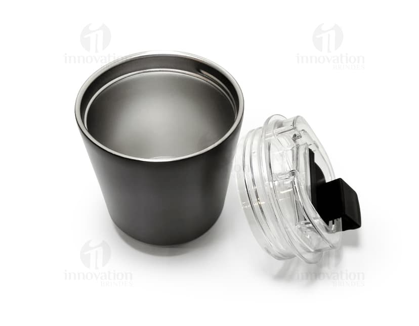 Copo térmico de 280ml em cerâmica preta e metal de cor prateada, com alça e tampa. O copo está vazio e posicionado sobre uma mesa de cozinha, ao lado de uma colher, uma jarra de café e alguns utensílios.