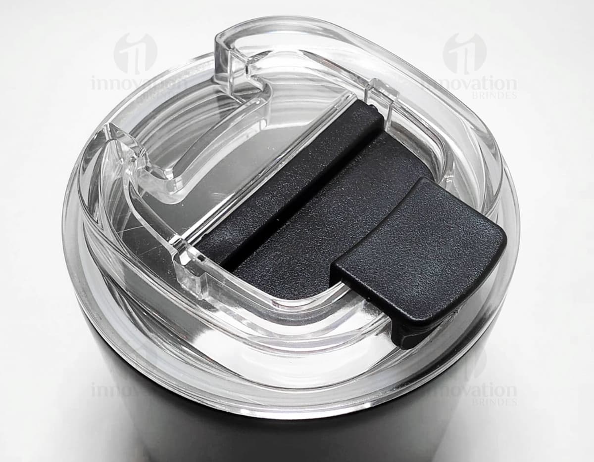 Copo térmico de 280ml em cerâmica preta e metal de cor prateada, com alça e tampa. O copo está vazio e posicionado sobre uma mesa de cozinha, ao lado de uma colher, uma jarra de café e alguns utensílios.