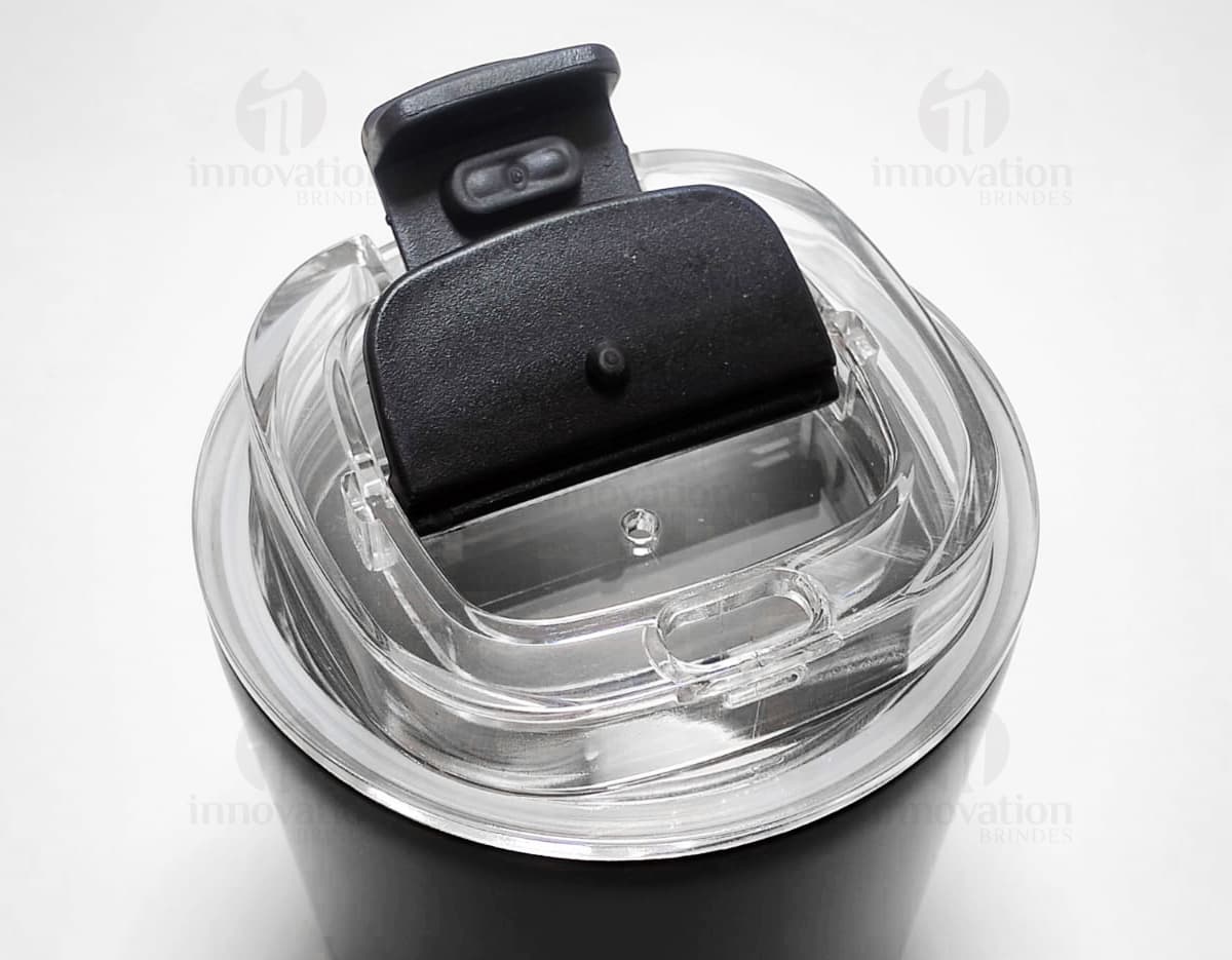 Copo térmico de 280ml em cerâmica preta e metal de cor prateada, com alça e tampa. O copo está vazio e posicionado sobre uma mesa de cozinha, ao lado de uma colher, uma jarra de café e alguns utensílios.