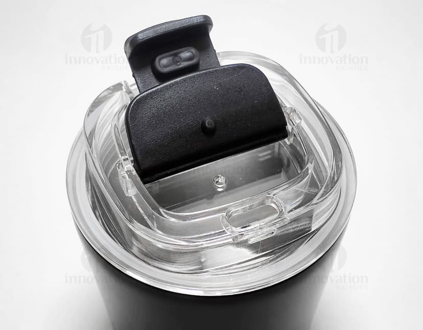 Copo térmico de 280ml em cerâmica preta e metal de cor prateada, com alça e tampa. O copo está vazio e posicionado sobre uma mesa de cozinha, ao lado de uma colher, uma jarra de café e alguns utensílios.