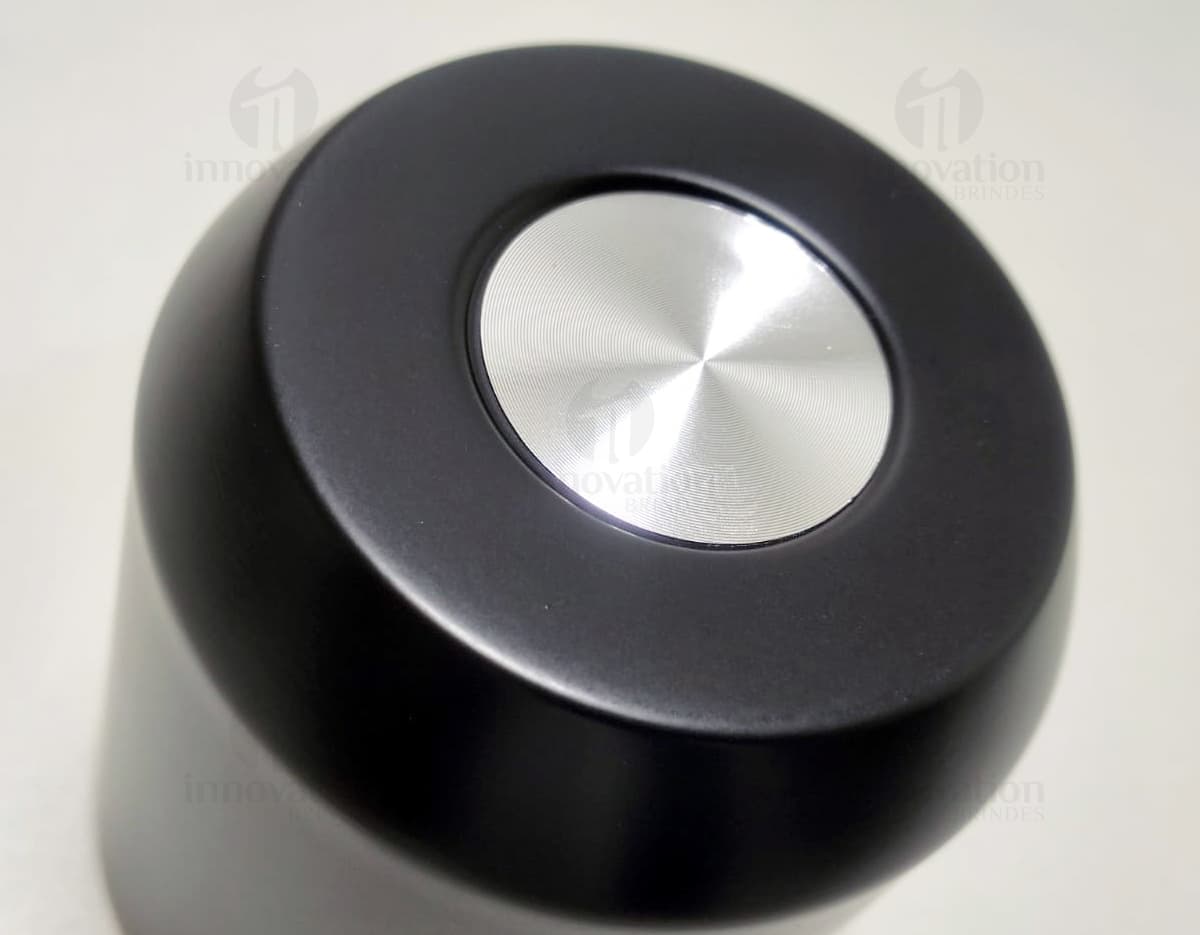 Copo térmico de 280ml em cerâmica preta e metal de cor prateada, com alça e tampa. O copo está vazio e posicionado sobre uma mesa de cozinha, ao lado de uma colher, uma jarra de café e alguns utensílios.