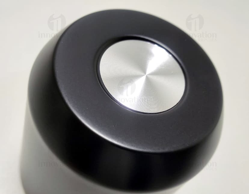 Copo térmico de 280ml em cerâmica preta e metal de cor prateada, com alça e tampa. O copo está vazio e posicionado sobre uma mesa de cozinha, ao lado de uma colher, uma jarra de café e alguns utensílios.