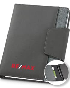 Notebook A5 com capa de couro sintético preto aberto sobre uma mesa de escritório. O caderno está vazio, com um teclado de computador ao lado, um estojo de negócios preto, um suporte para papel, discos magnéticos e notas espalhadas.