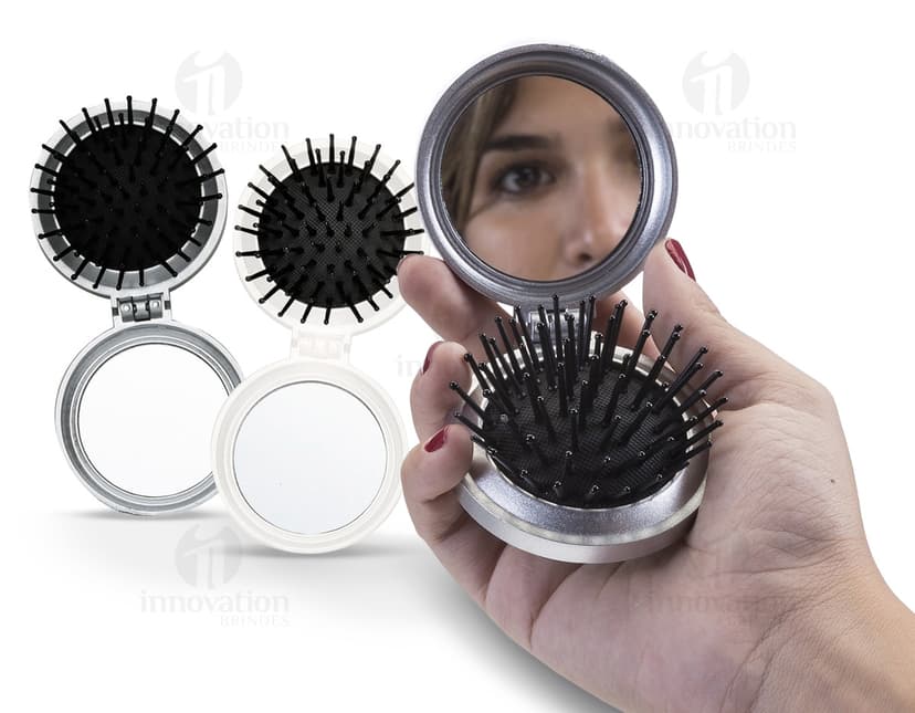 Espelho de bolsa com escova preta e vermelha, feito de vidro e metal, com um botão redondo para fechar. Possui um ícone de lupa prateada, símbolo de pesquisa e ampliação. O objeto é um instrumento de maquiagem compacto e prático para levar na bolsa, ideal para retoques durante o dia.