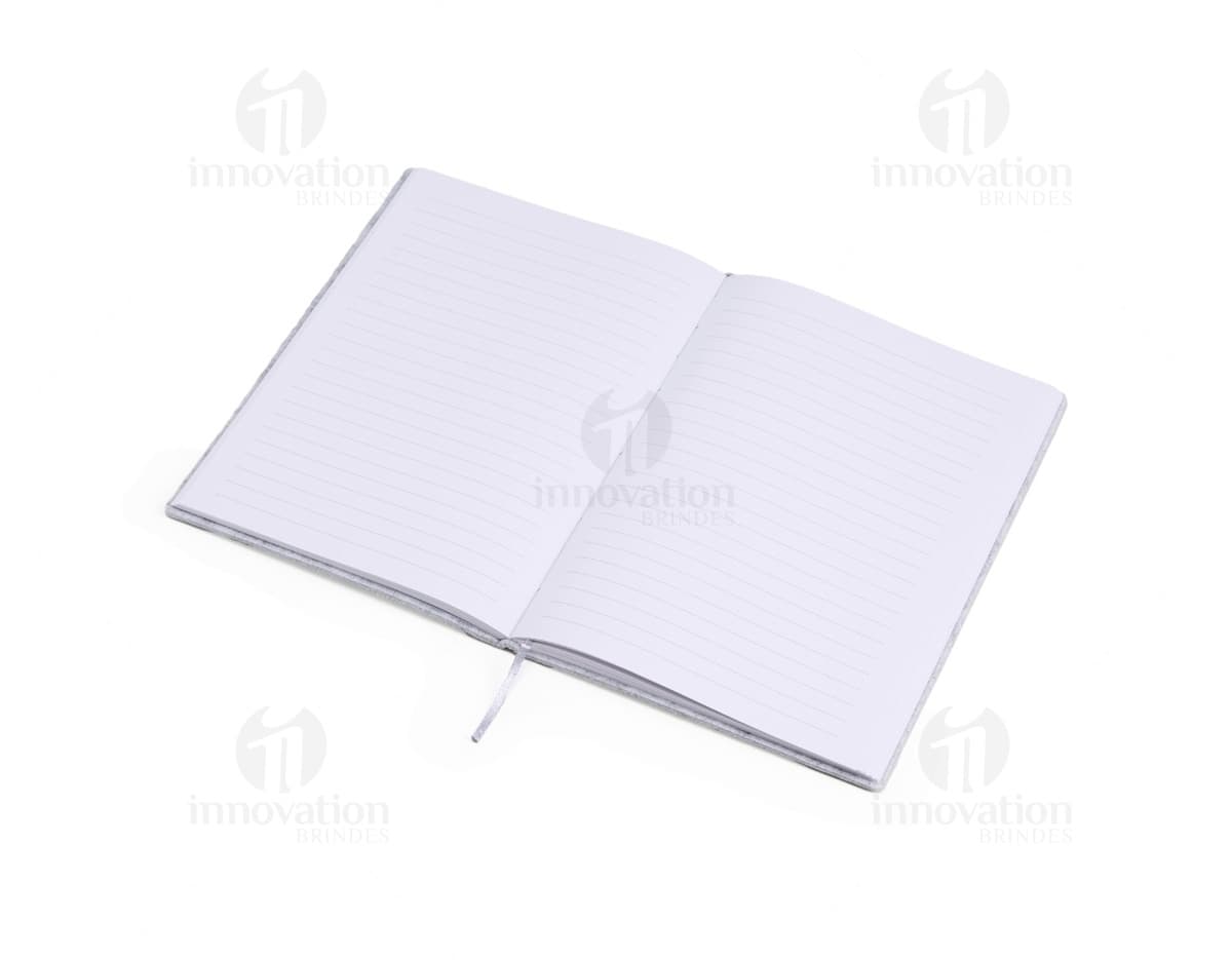 caderno A5 de feltro preto, sem pauta, aberto em uma página em branco. Ao lado, um notebook e um envelope preto. No fundo, livros e objetos de escritório.