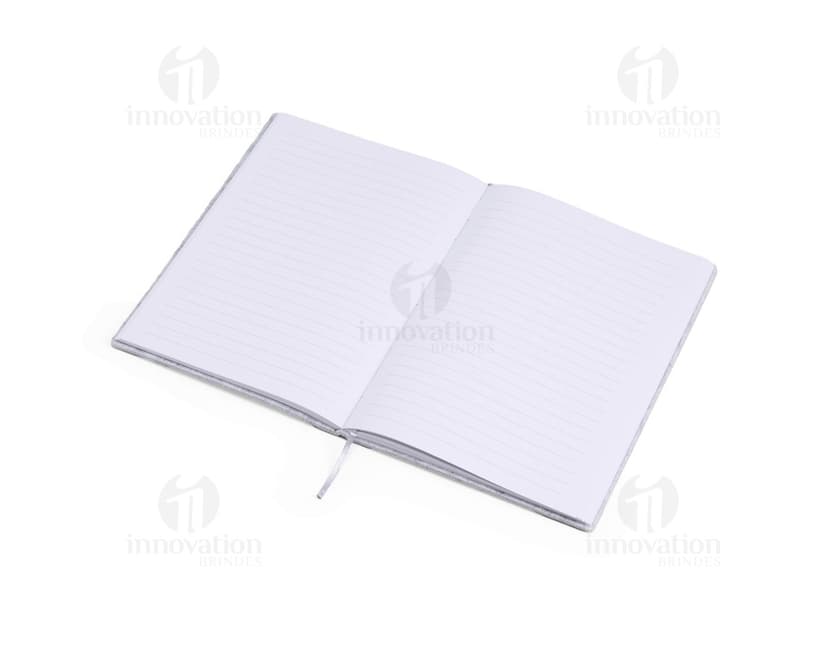 caderno A5 de feltro preto, sem pauta, aberto em uma página em branco. Ao lado, um notebook e um envelope preto. No fundo, livros e objetos de escritório.